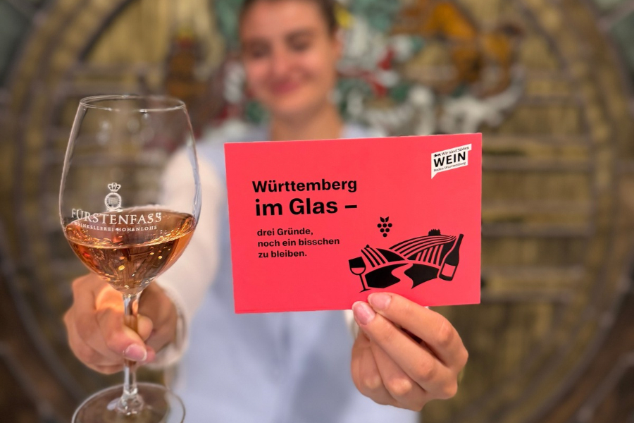 Eine Person hält ein Fürstenfass-Weinglas mit Rosé und eine Karte zur Aktion „Württemberg im Glas“ in die Kamera.